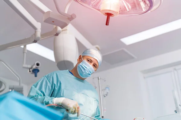 Confident surgeon wearing protective mask looking at the patient and feeling concentrated during the operation. Medicine concept. Low angle