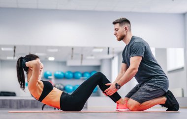 Spor salonundaki minderde koçunun yardımıyla karın egzersizi yapan genç bir kadın. Kas çalıştırıcı çekici dişinin karın kaslarını çalıştırmasına yardım eder