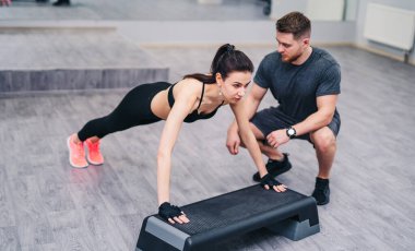 Çapraz formda bir kadın spor salonundaki özel bir bankta elleriyle şınav çekiyor. Koyu renk saçlı, spor hocası olan bir kadın. Yakın plan.