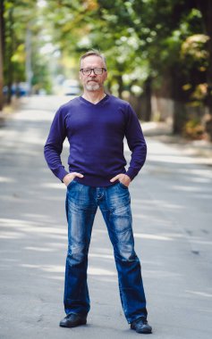 Açık havada gri saçlı, yakışıklı, orta yaşlı bir adam. Mavi kazak. Gözlük. Sakallı, kendine güvenen iş adamı. Arka plan bulanık. Ellerini cebine koy.