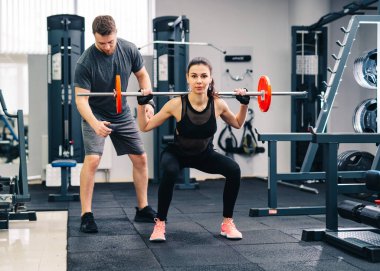 Spor, vücut geliştirme, yaşam tarzı ve insan konsepti. Genç adam ve kadın halterle kaslarını esnetiyor ve spor salonunda omuz presi yapıyor. Antrenör ve fitness kızı spor salonunda antrenman yapıyor.