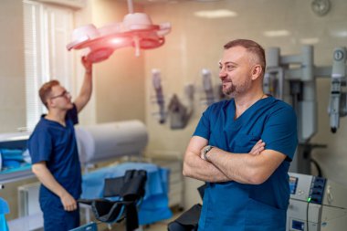Erkek cerrah koruyucu giysiler giyip arka planda meslektaşıyla dikiliyor ve başka tarafa bakıyor. Operasyon sırasında sıhhiye. Tıp konsepti