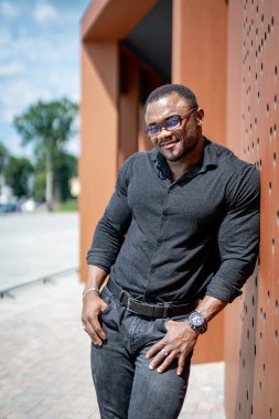 Binada duran, duvara yaslanan yakışıklı, sportif adam. Güneşin aydınlattığı koyu renk elbiseli, gülümseyen bir adam. Sokakta duran gözlüklü Afrikalı adam..