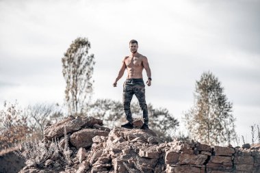 Kamuflaj pantolonlu güçlü yakışıklı vücut geliştirici kayanın üzerinde duruyor. Uçurumun tepesinde tişörtsüz gülümseyen kaslı adam. Gömleksiz beyaz adam kayanın üzerinde güneş ışığının altında.