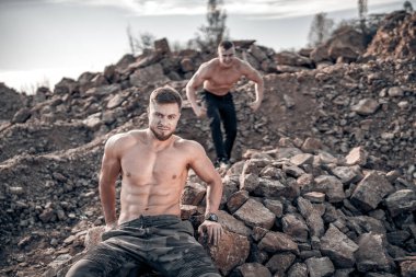 Güçlü kasları olan gömleksiz iki atletik adam. Kayalıklarda oturan sakallı adam. Büyük taşların arkasında güçlü ve zinde erkekler.