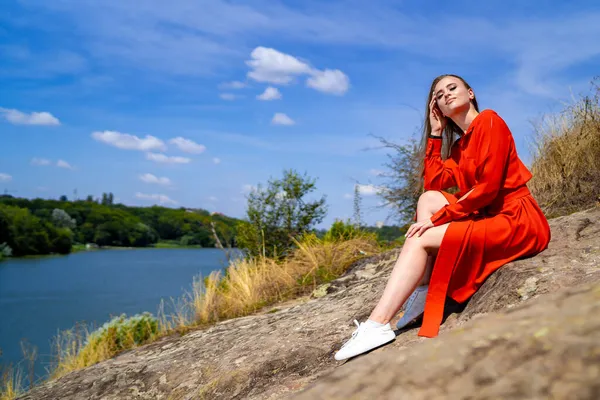 自然を背景にした赤いロングドレスの美しい若い女性 プロの夏の写真撮影 閉鎖だ 少女は岩の上に座って 長い美しい足 — ストック写真