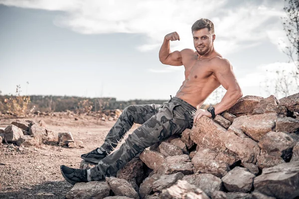 Beyaz tenli yakışıklı erkek güçlü pazılarını sağ elinde sergiliyor. Sağlıklı üstsüz, yarı çıplak, kayalıklarda yatan bir adam. Gülümseyen, kamuflaj pantolonlu, uyumlu bir erkek..