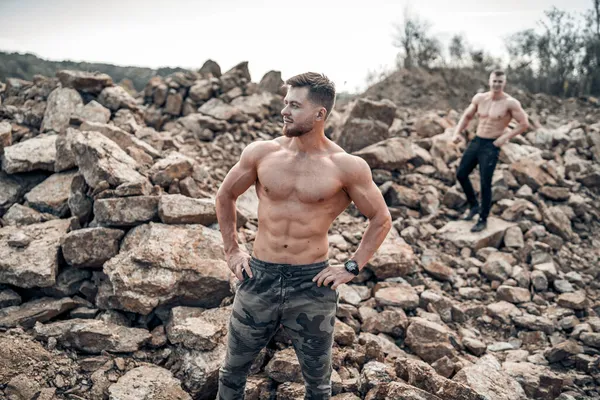 Yakışıklı sporcular formda ve kaslı vücutlarını sergiliyorlar. Erkekler elleri kalçalarında durur. Kayalık tepelerin arasında iki üstsüz beyaz erkek..