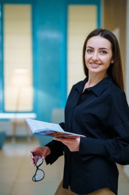 Çekici, gözlüklü, gülümseyen ve kameranın önünde kitap okuyan güzel bir kız. Açık renk iç renkler. Yakın çekim.