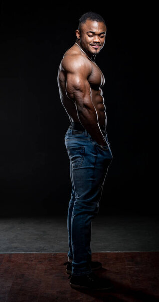 Very muscular afro american man with naked torso poses on black background. Bodybuilder in jeans looking at the camera