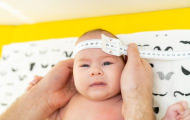 Yeni doğan bebeği muayene eden bir çocuk doktorunun fotoğrafı. Doktor, bebeğin kafa büyüklüğünü ölçmek için mezura kullanıyor. Yakın çekim.
