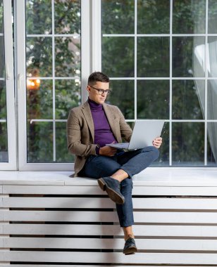 Takım elbiseli genç bir erkek oturuyor ve ekrana bakıyor. Pencerenin kenarında oturan yakışıklı bir iş adamı ve dizüstü bilgisayarın başında çalışıyor.