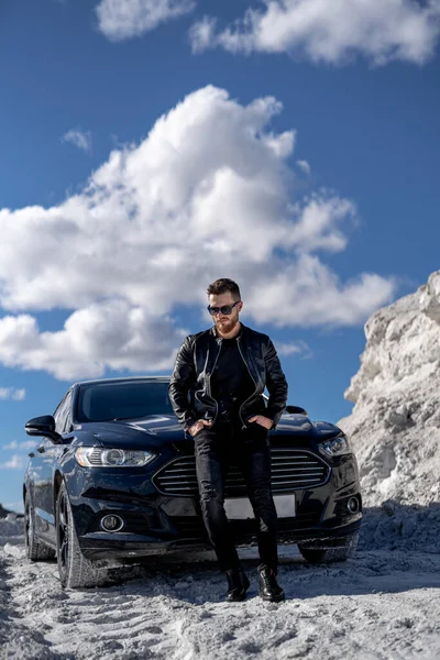 一个穿着时髦太阳镜和皮夹克的时髦大胡子男人站在黑色汽车旁边的画像 白色岩石背景的时装肖像 — 图库照片