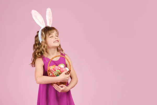 Flicka i påsk kanin öron håller i händerna en korg med ägg på rosa bakgrund — Stockfoto