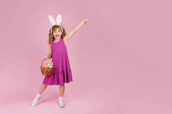 Flicka i påsk kanin öron håller i händerna en korg med ägg på rosa bakgrund — Stockfoto