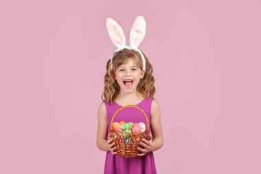 Paskalya tavşanı kulaklı kız pembe arka planda yumurtalarla dolu bir sepeti elinde tutuyor.