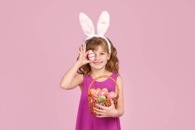 Paskalya tavşanı kulaklı kız pembe arka planda yumurtalarla dolu bir sepeti elinde tutuyor.