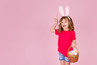 Paskalya tavşanı kulaklı kız pembe arka planda yumurtalarla dolu bir sepeti elinde tutuyor.