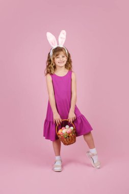 Paskalya tavşanı kulaklı kız pembe arka planda yumurtalarla dolu bir sepeti elinde tutuyor.