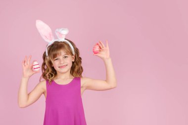 Paskalya tavşanı kulaklı küçük kız ellerinde renkli yumurtalar tutarken gülümsüyor. 