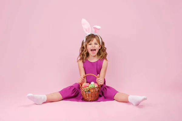 Fille dans les oreilles de lapin de Pâques tient dans les mains un panier avec des œufs sur fond rose Image En Vente