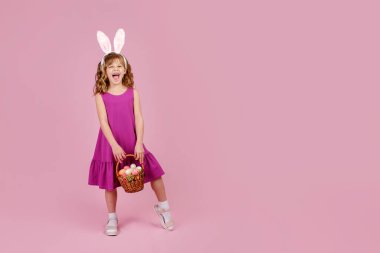 Paskalya tavşanı kulaklı kız pembe arka planda yumurtalarla dolu bir sepeti elinde tutuyor.