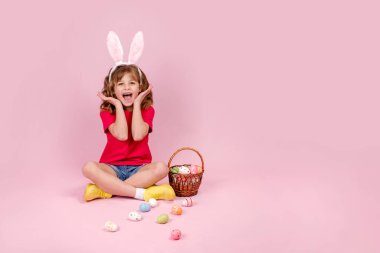 Paskalya tavşanı kulaklıklarıyla gülümseyen küçük güzel kız Paskalya yumurtasını elinde tutuyor, pembe arka planda dağınık renkli yumurtalarla sepetin yanında oturuyor, 