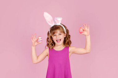 Paskalya tavşanı kulaklı küçük kız ellerinde renkli yumurtalar tutarken gülümsüyor. 