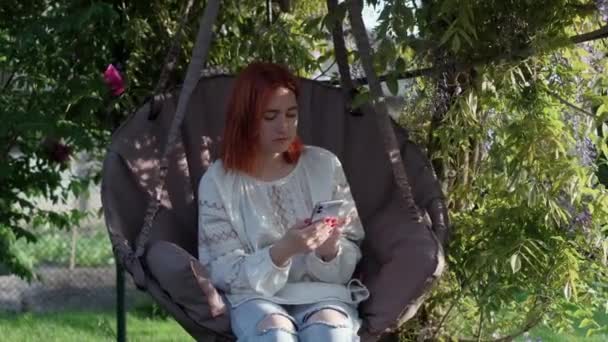 Ung flicka använder smartphone och gunga, sitter i hängande stol på bakgården — Stockvideo