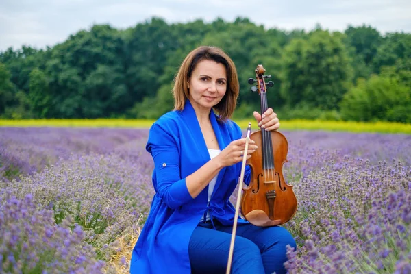 Full body adult woman violinist playing violin and sitting on summer lavender field, romantic musician in blue dress enjoying walking on nature