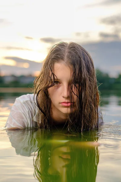 Face of young beautiful teenage girl in a white shirt sits in the water of a lake, while swimming and looks directly into the camera at sunset
