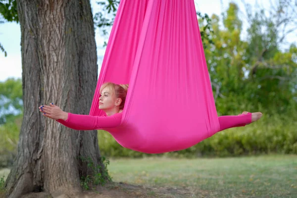 美しい若い女性は公園の木にかかってピンクのハンモックに横たわっている間 空中ヨガを練習します 女性は屋外で反重力ヨガを練習します — ストック写真