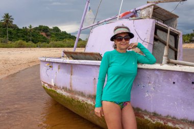 Trancoso, Bahia, Brezilya 'daki Praia dos Nativos' ta eski bir terk edilmiş ve yarı batık bir balıkçı teknesinin yanında poz veren kadın.