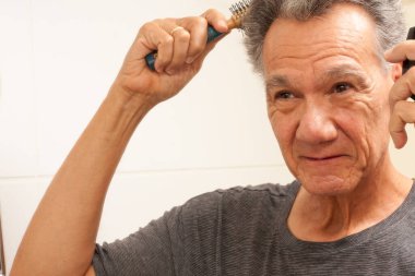 Mature Man 60plus combing his mostly gray hair in the back of his head in front of a mirror