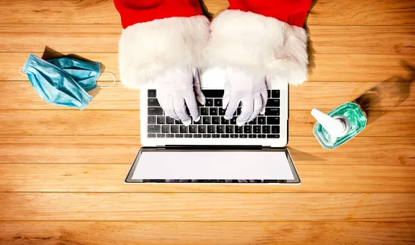 Papai Noel usando um computador portátil com uma máscara e uma garrafa de desinfetante — Fotografia de Stock