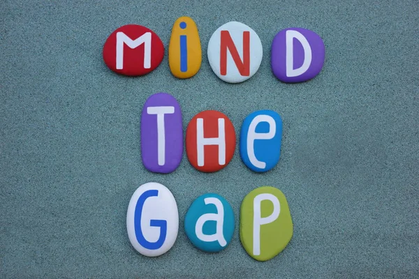 Mind the gap, creative logo composed with multi colored stone letters ovver green sand