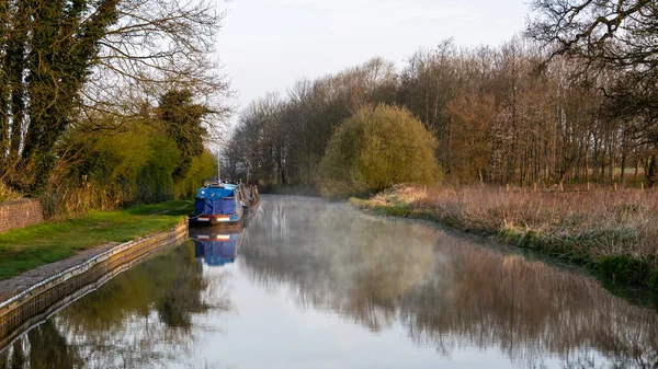 A peaceful tranquil scene looking down a canal. The water is calm and there are reflections together with an early morning mist rising
