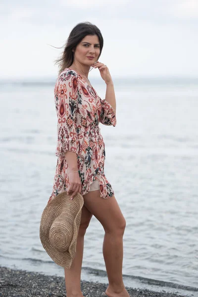Модная белая женщина, держащая шляпу на пляже. Девушка улыбается на побережье. Летнее время в стиле жизни — стоковое фото