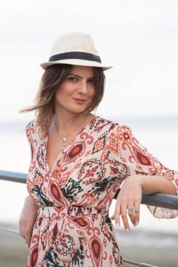 Yazlık elbise giyen ve şapkalı bir kadın kameraya bakıp gülümsüyor. Plajda yaz konsepti