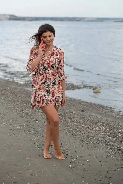 Улыбающаяся юная брюнетка-кавказка болтает по красному мобильному телефону на пляже. Люди образ жизни — стоковое фото