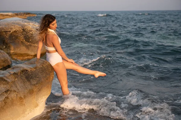 Mujer joven con traje de baño blanco jugando en el agua en la playa al atardecer — Foto de Stock