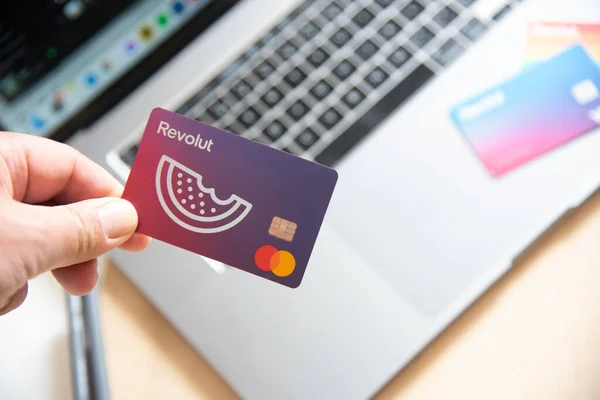 Human hand holding a revolut debit card. Mobile phone app digital payments online with laptop computer — 스톡 사진