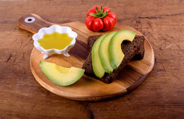Olive oil in white cup, red tomato, rye bread with avocado on wood cutting board.