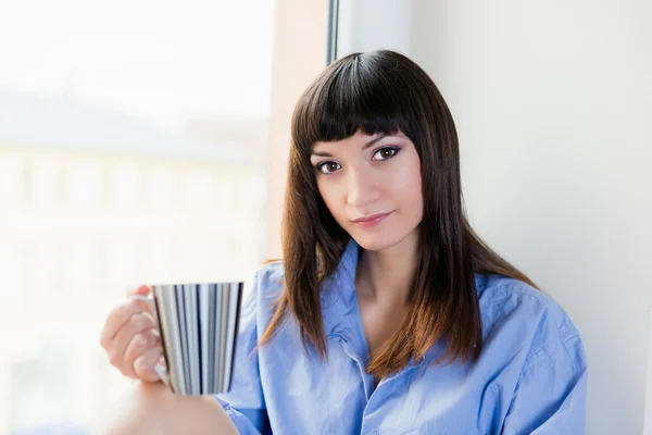 Krásná brunetka v modré košili pití mu první ranní šálek kávy nebo čaje — Stock fotografie