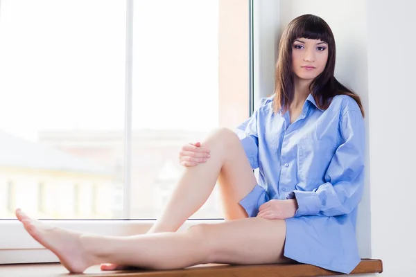 Beautiful young woman in blue shirt sitting on the window sill — Stock Photo, Image