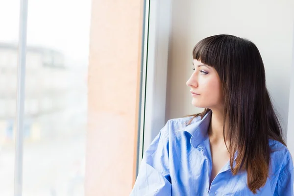 Mujer con camisa azul mirando por la ventana — Foto de Stock