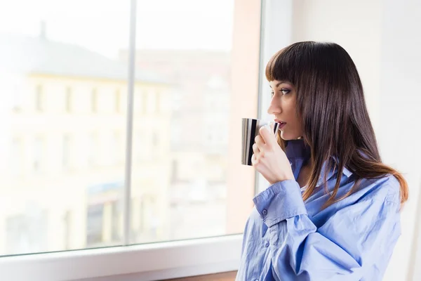 Женщина, стоящая у окна и пьющая кофе — стоковое фото