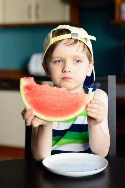 Niño de 4 años con gorra come una sandía en la mesa de la cocina —  Fotos de Stock