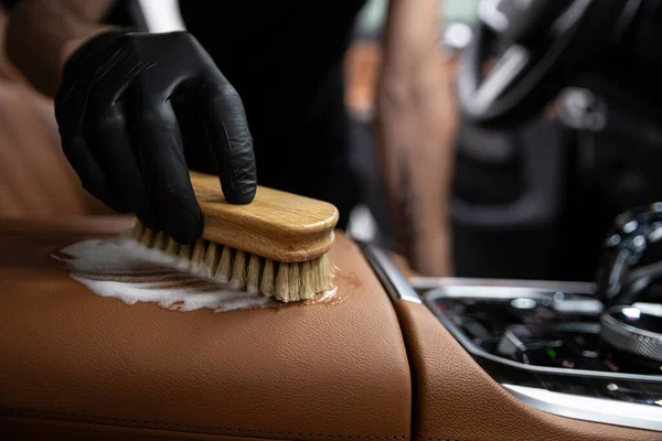 car detailing studio employee cleans the brown leather upholstery of a car with a detergent and a leather cleaning brush