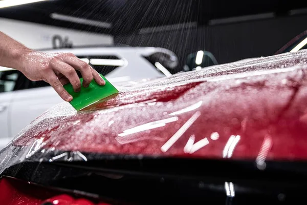 Man car detailing studio employee applies a colorless protective film to the body of a red car